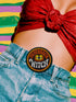 A round, black embroidered patch with a thick, textured merrow border stitched in alternating orange and yellow thread. In the center, the words "PAPERBACK WITCH" are embroidered in a retro 70s-style font. The word "PAPERBACK" is arched across the top in bold orange thread with a thin yellow outline. The word "WITCH" is across the bottom in puffy, bright white thread with a thin yellow outline. Between the words is a central icon of an open book, containing a stylized all-seeing eye.